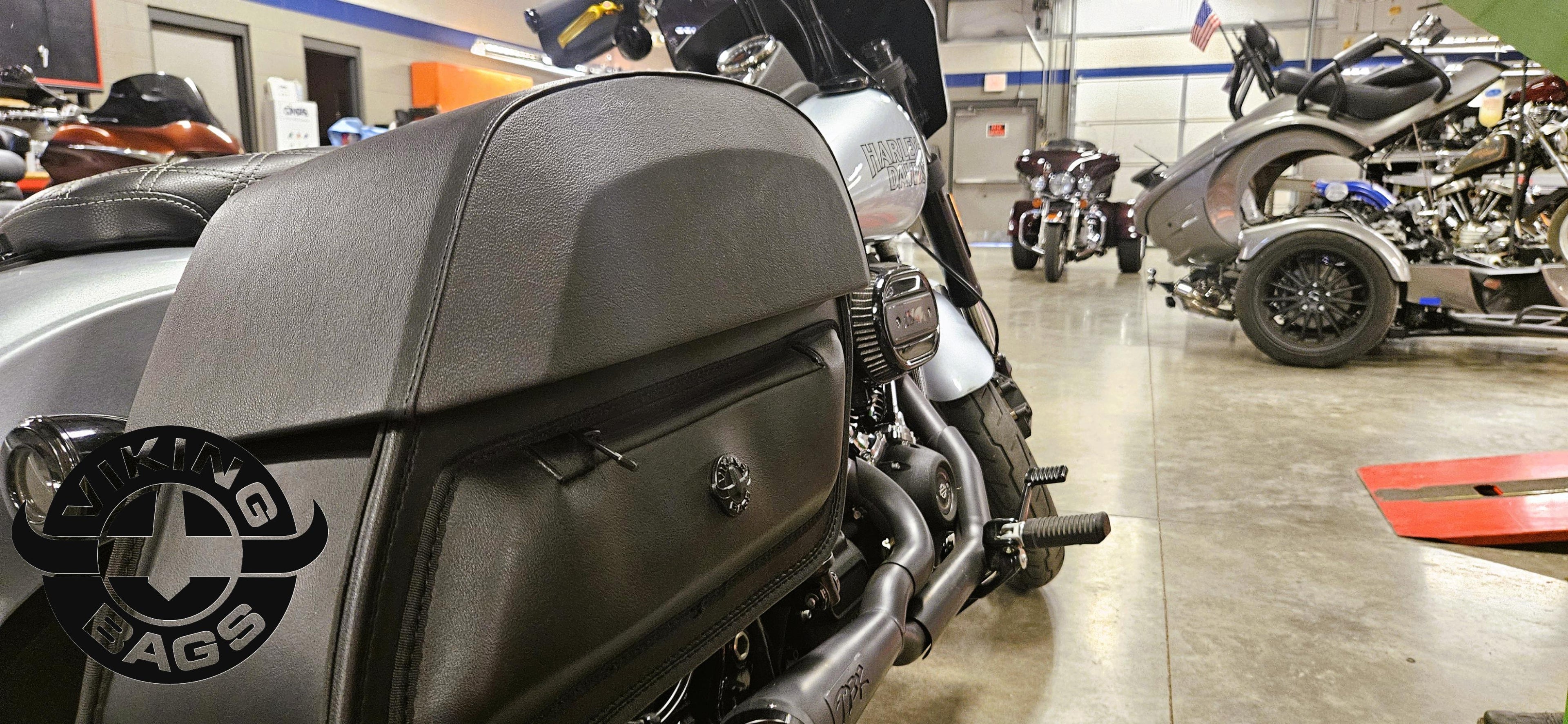 Close-up of a motorcycle saddlebag with a blurred background of a garage with multiple motorcycles.
